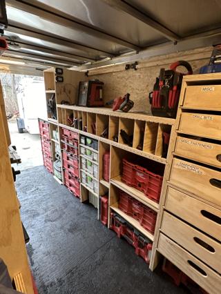 View down the length of the trailer showing shop-made plywood drawers on the right and open shelving holding Milwaukee Packout crates and Festool Systainers side by side