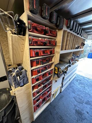 Trailer shelving with a tall bank of Milwaukee Packout organizers hand-labelled Spray Touchup and Kreg Screws, with batteries mounted along the shelf edge