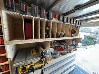 Lower trailer shelf holding a DeWalt jobsite table saw and two mitre saws, with guide rails stored beneath and vertical plywood dividers above