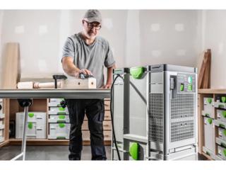 A craftsman working a piece of timber at a bench with a Festool SYS-AIR running beside him, shelves of Systainers behind