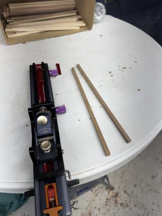 The Chopstick Master jig on a work table beside a finished pair of chopsticks, with a box of square blanks behind
