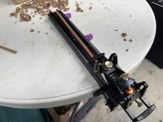 A chopstick blank clamped in the Chopstick Master track with the block plane at the end of its run and shavings scattered across the table