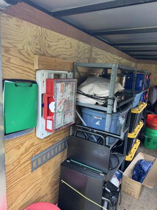 Light grey Systainer³ Base screwed to the plywood wall of a cargo trailer with a red SYS-Aid M 89 Organizer clipped into it, its clear lid showing compartments of first aid supplies, a green clipboard hanging alongside and steel shelving with totes and buckets to the right