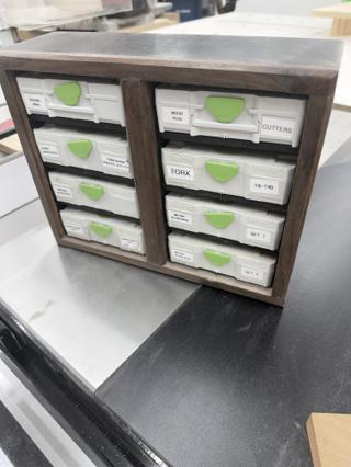 Dark stained wooden cabinet on a table saw wing holding eight light grey Systainer³ XXS cases in two columns, each with a green latch and a printed white label reading SQUARE DRIVE, WOOD PLUG CUTTERS, TORX and similar