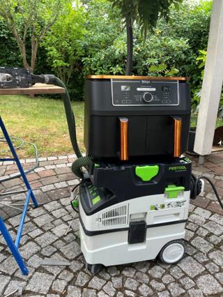 A black Ninja dual-zone air fryer with copper trim sitting squarely on the systainer-format lid of a Festool CLEANTEC dust extractor, hose attached, on a paver patio in front of a hedge