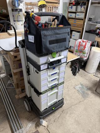 The same stack from the front with the SYS-Roll handle extended, showing the TANOS Sortainer with blue drawer pulls above a Festool Sortainer with green pulls, and the work surface folded out on the right