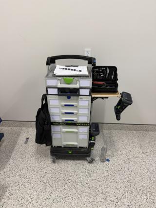 The stack on a speckled floor against a white wall, the work surface folded out and holding an open bit case, a cordless drill hanging from the side below it and a black bag hanging from the other side