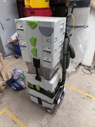 Three Festool systainers stacked on a Festool dust extractor in a shop, with the hose coiled and hanging from the side of the stack