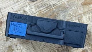 A closed anthracite Systainer³ XXS on a workbench, TANOS and Systainer³ molded into the lid, with FIRST AID hand-written on blue tape in the label recess