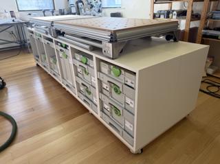 A long island of white SYS-CAB-M-AZ cabinets on casters in a studio with a hardwood floor, two Festool MFT tops resting on it end to end; the nearest bays hold Festool Systainer³ cases in SYS-AZ drawers, and a few bays at the far end are still empty