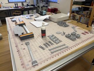 A Festool MFT top covered with a gridded cutting mat, with gray 3D-printed suspension arms, shock bodies and chassis blocks for an RC car laid out next to an orange combination square, a bottle of adhesive and an open notebook