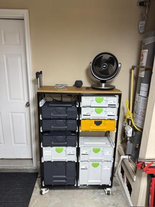A two-bay rack of black 20×20 aluminum extrusion on casters in a garage, eleven systainers sliding on white Systainer³ Rails in two columns, three anthracite and one Daffodil Yellow among the Festool boxes, a wood worktop on top with a fan on it, and a track saw guide rail standing on the left side